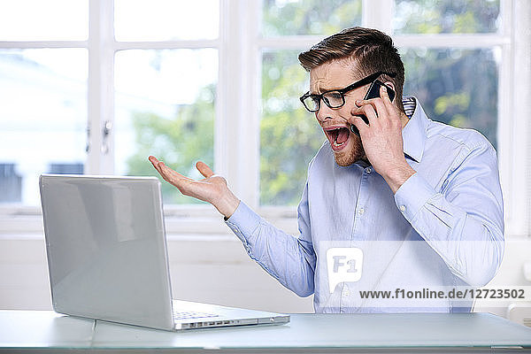 Mann in blauem Hemd; Brille; Bart; ernst; Fenster im Hintergrund unscharf; sitzend; Blick auf seinen Computer-Laptop; Telefon am Ohr  Hand in der Luft  schreiend .