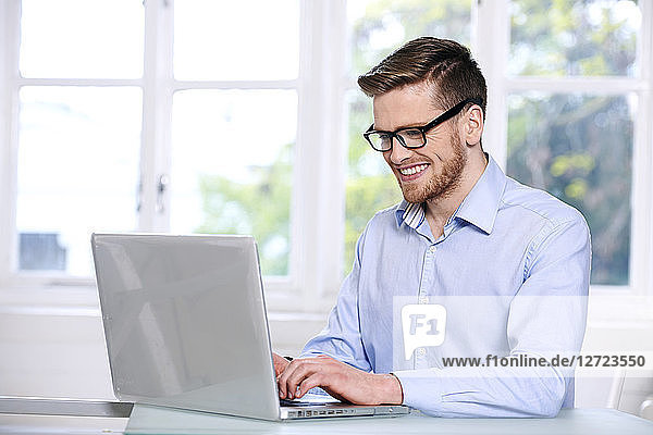 Mann in blauem Hemd  Brille  Bart  lächelnd  Fenster im Hintergrund unscharf  sitzend  tippt auf einem Computer-Laptop