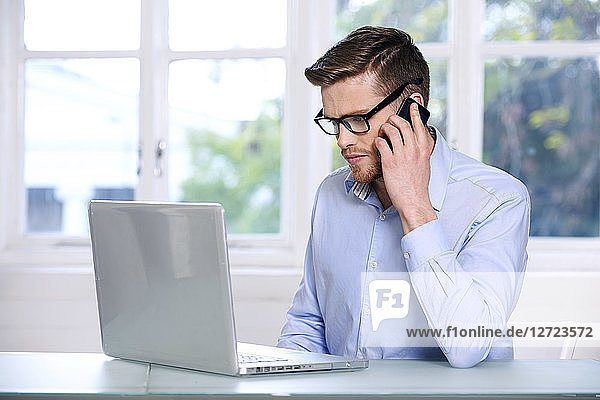 Mann in blauem Hemd; Brille; Bart; ernst; Fenster im Hintergrund unscharf; sitzend; Blick auf seinen Computer-Laptop; Telefon am Ohr'.