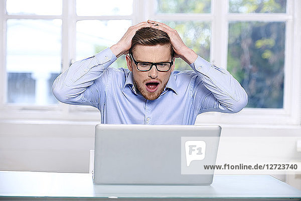 Mann in blauem Hemd  Brille  Bart  ernst  Fenster im Hintergrund unscharf  Mund geöffnet; Hände auf dem Kopf; sitzend  auf seinen Laptop blickend .