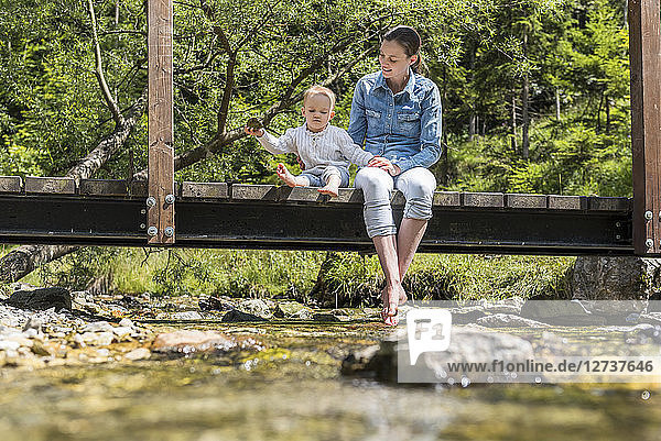 Mother and daughter sitting on wooden bridge girl throwing stone into mountain stream