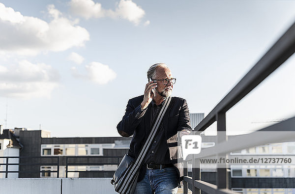 Mature man standing on rooftop  leaning on railing  using smartphone