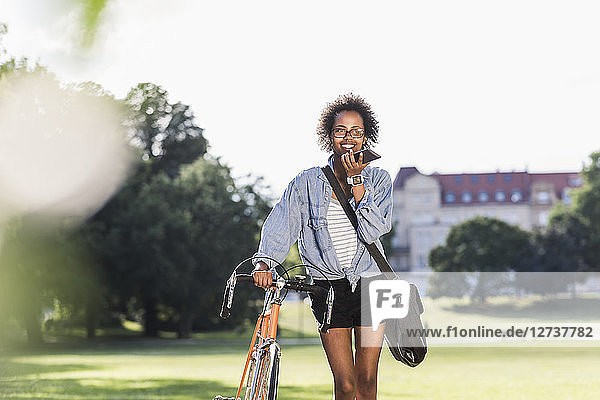Smiling young woman with cell phone and bicycle in park