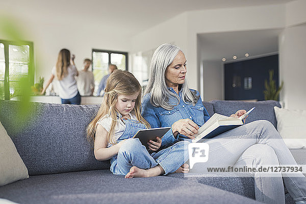 Grandmother and granddaughter sitting on couch  reading together book and tablet pc