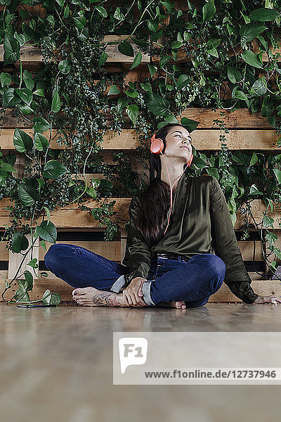 Young woman sitting on floor at wall with climbing plants listening to music