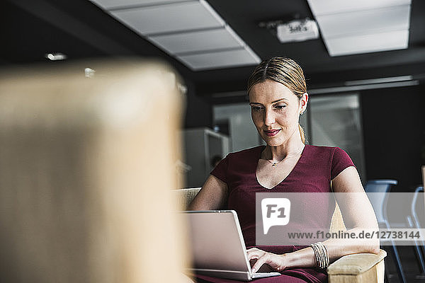 Businesswoman in office lounge wearing burgundy dress using laptop
