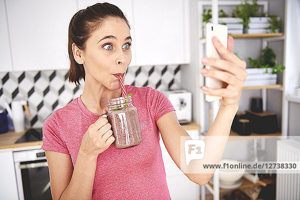 Portrait of young woman taking selfie with smartphone in the kitchen while drinking smoothie