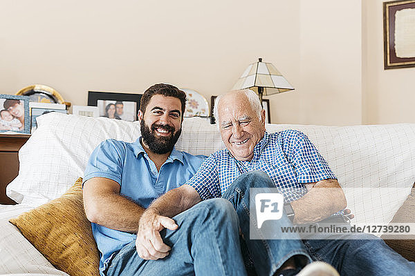 Portrait of adult grandson and his grandfather sitting on the couch at home tickling each other