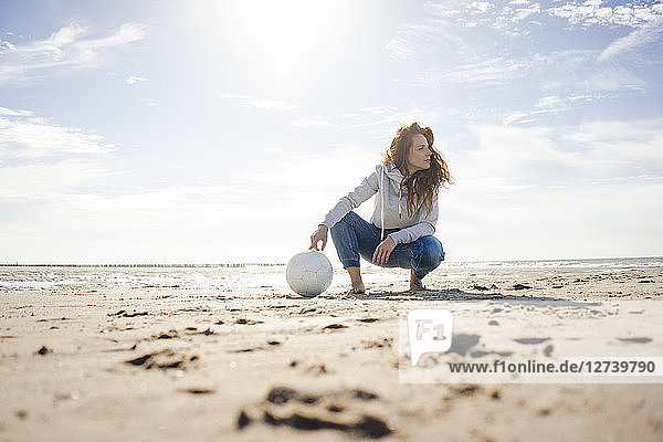 Woman with soccer ball crouching on the beach  looking away