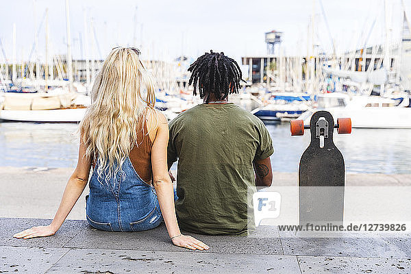 Spain  Barcelona  back view of multicultural young couple sitting side by side with longboard at marina