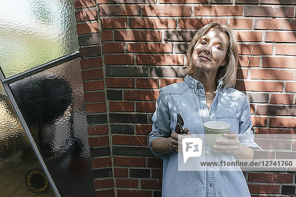 Relaxed mature woman leaning at house wall holding cup and pruner