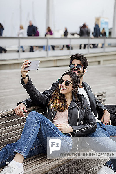 Spain  Barcelona  happy young couple resting on a bench taking a selfie