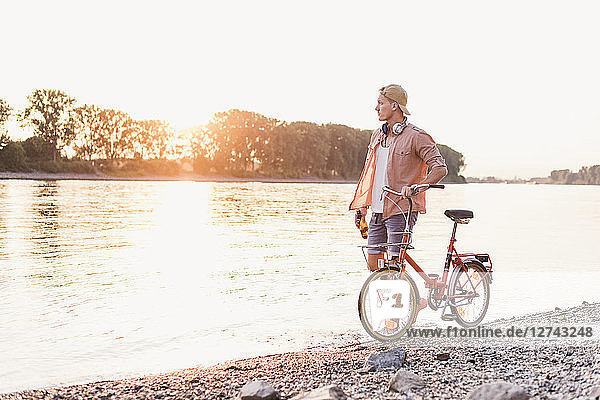 Young man walking with bicycle at the riverbank at sunset