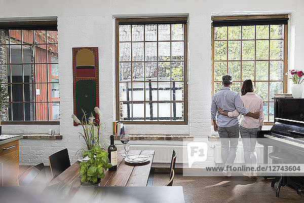 Mature couple standing in their comfortable loft  looking out of window  rear view