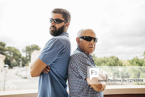 Portrait of adult grandson and his grandfather wearing sunglasses standing back to back on balcony