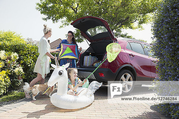 Lesbian couple and daughter loading kite and inflatable unicorn into car hatchback in sunny driveway