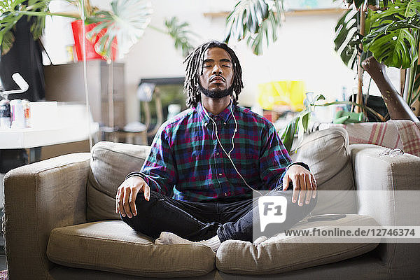 Serene young man meditating with headphones on apartment sofa
