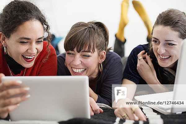 Laughing young women friends hanging out  enjoying digital tablet and laptop on bed