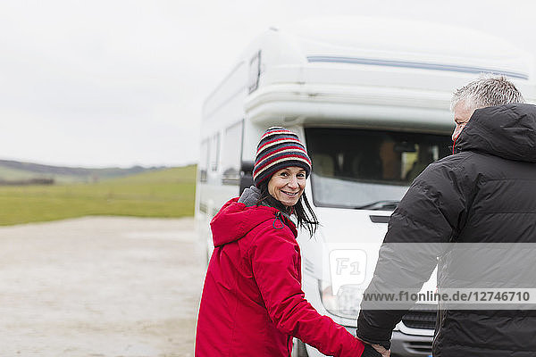 Porträt eines lächelnden Paares in warmer Kleidung  das sich vor einem Wohnmobil an den Händen hält