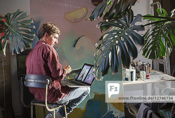 Männlicher Künstler malt und benutzt einen Laptop in einem Kunststudio
