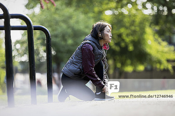 Active senior female runner with headphones tying shoe in park