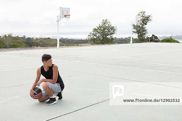 Männlicher jugendlicher Basketballspieler hockt mit dem Ball auf dem Basketballfeld