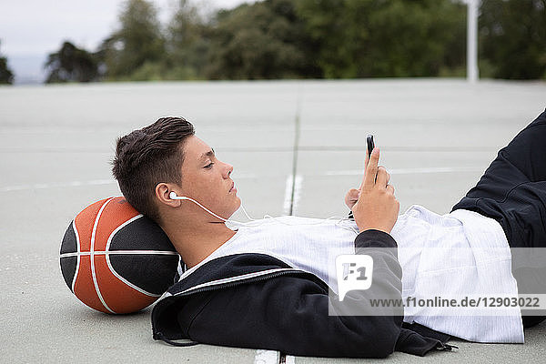 Männlicher jugendlicher Basketballspieler liegt auf Basketballfeld und schaut auf Smartphone
