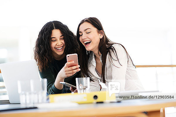 Teenage schoolgirls at classroom desk laughing at smartphone