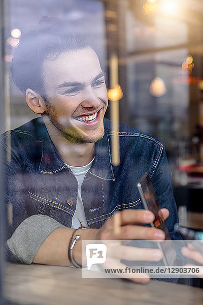 Männlicher Student zeigt Smartphone am Fensterplatz eines Cafés  Blick durchs Fenster