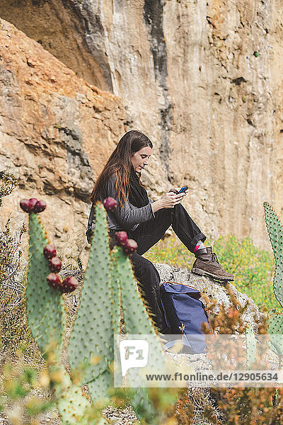 Spain  Alquezar  young woman sitting on a rock using smartphone
