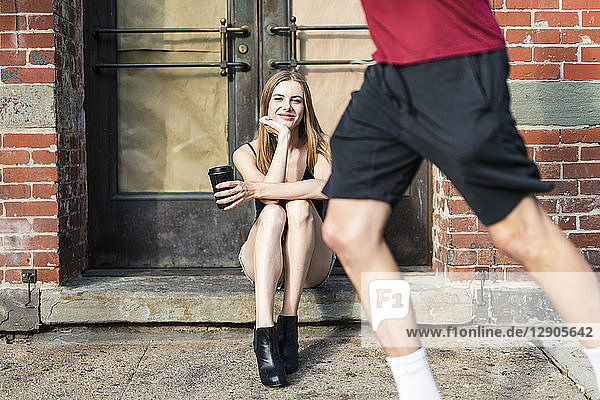 Young woman sitting in the street on a doorstep  holding a cup of coffee  jogger passing