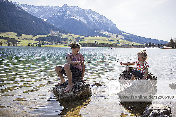 Austria  Tyrol  Walchsee  brother and sister sitting on boulders in the lake playing with sticks
