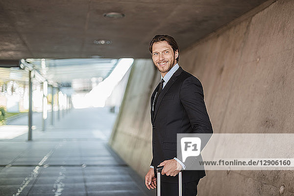 Smiling businessman with rolling suitcase standing in underpass