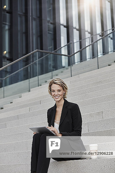 Portrait of smiling businesswoman with tablet and coffee to go sitting on stairs in front of modern office building