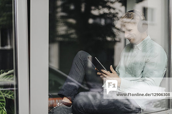 Young man sitting at the window  listening music with headphones and digital tablet
