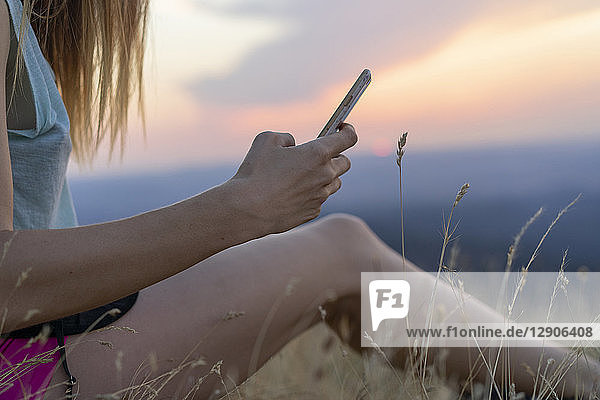 Close-up of young woman using cell phone sitting in grass during sunset