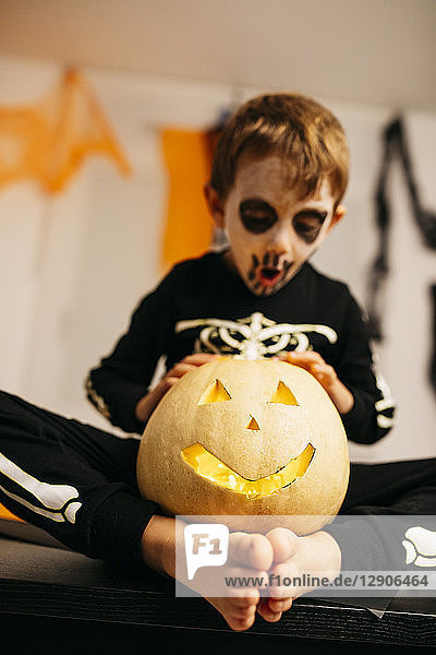 Little boy with painted face and fancy dress sitting barefoot on table with Jack O'Lantern