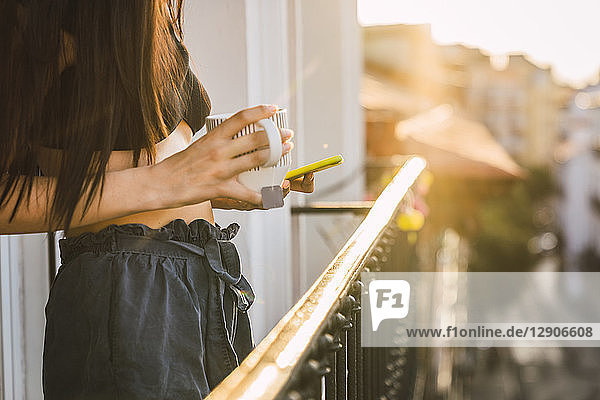 Close-up of young woman on balcony above the city at sunset holding cell phone and cup of tea