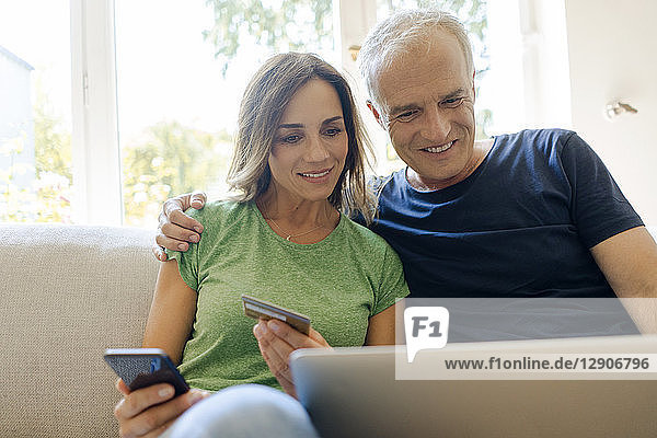 Smiling mature couple sitting on couch at home shopping online with laptopand smartphone