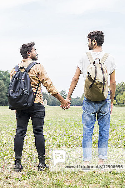 Back view of young gay couple with backpacks standing hand in hand on a meadow