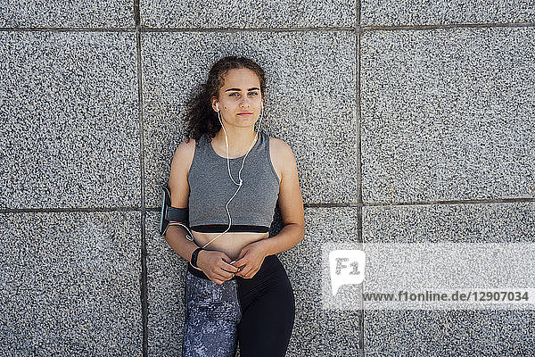 Portrait of young athletic woman wearing earbuds leaning against a wall