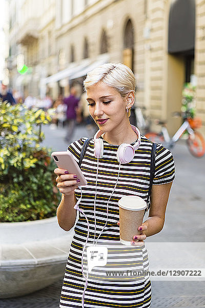 Portrait of young woman wearing striped dress standing on street with coffee to go looking at cell phone