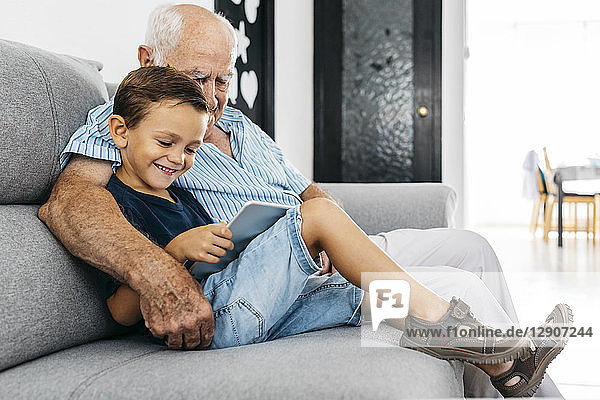 Grandfather and grandson sitting together on the couch at home looking at digital tablet