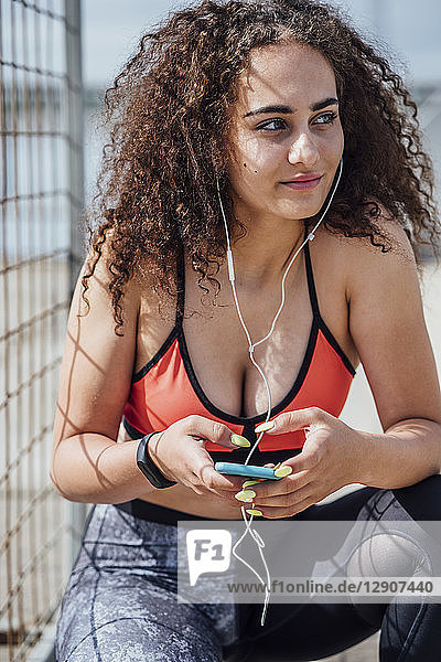 Smiling young athletic woman with smartphone and earbuds crouching outdoors
