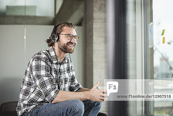 Happy casual businessman with cup of coffee and headset in office