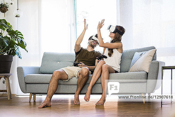 Happy couple sitting on couch at home wearing VR glasses high fiving