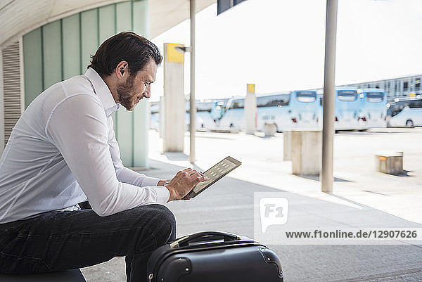 Smiling businessman with suitcase sitting at bus terminal using tablet