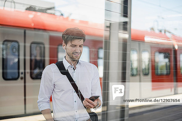 Smiling businessman on station platform with earphones and cell phone