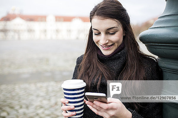Portrait of young woman with coffee to go leaning against lamp pole looking at cell phone