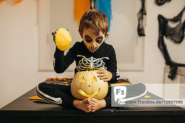 Portrait of little boy with painted face and fancy dress sitting on table looking inside of Jack O'Lantern
